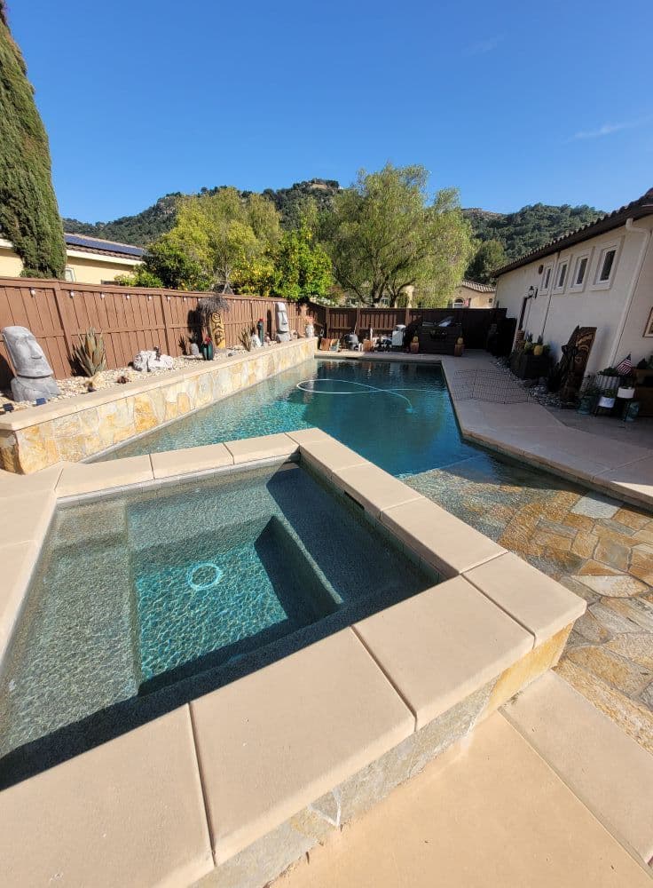Beautiful backyard pool and spa surrounded by stone, with lush greenery and a clear blue sky.
