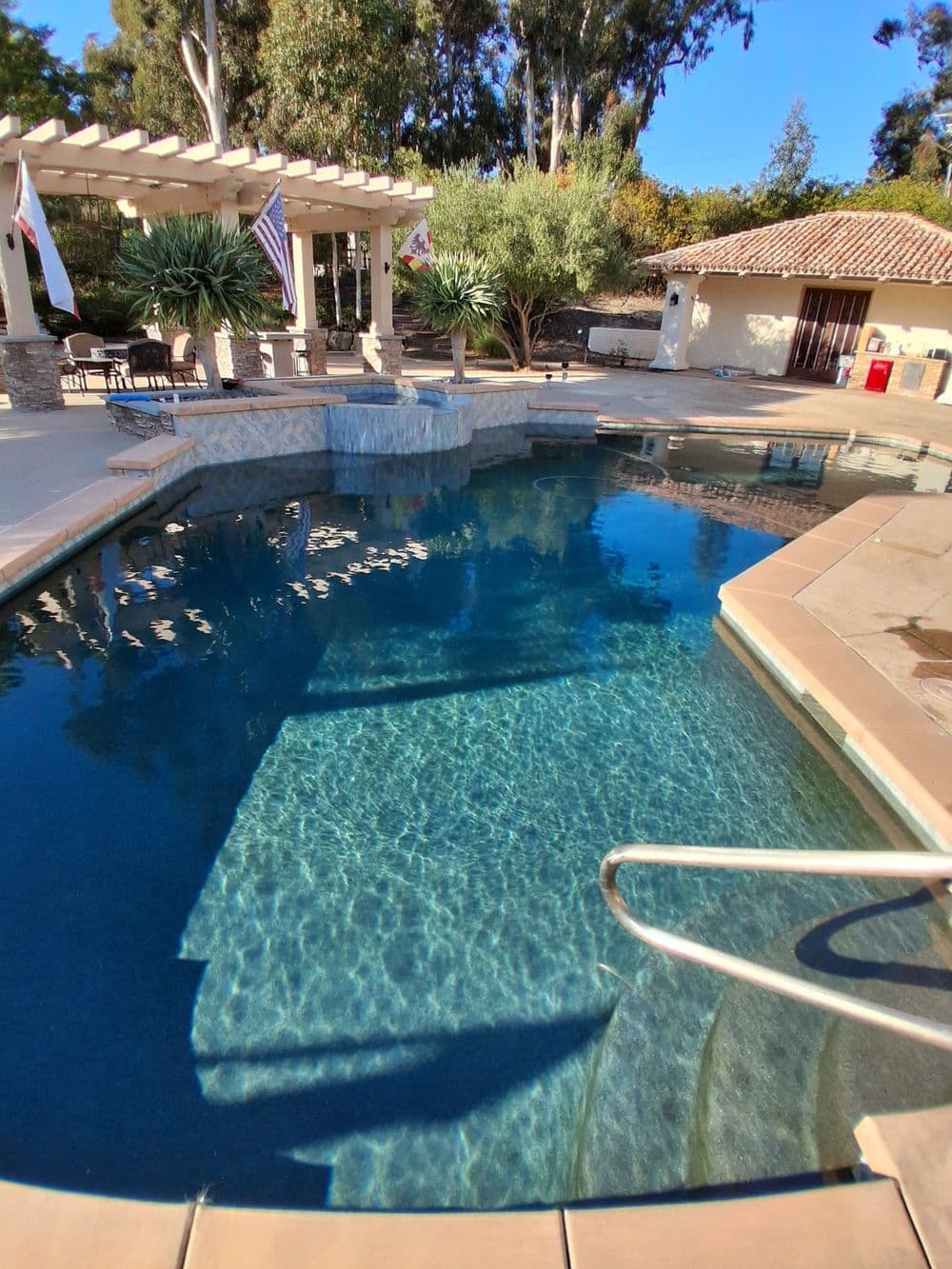 sparkling swimming pool with sunlit water, surrounded by lush greenery and outdoor seating areas