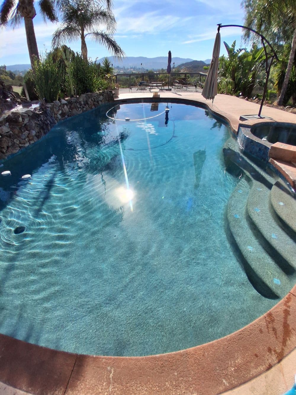 Luxury swimming pool with clear water, palm trees, and mountain views on a sunny day.